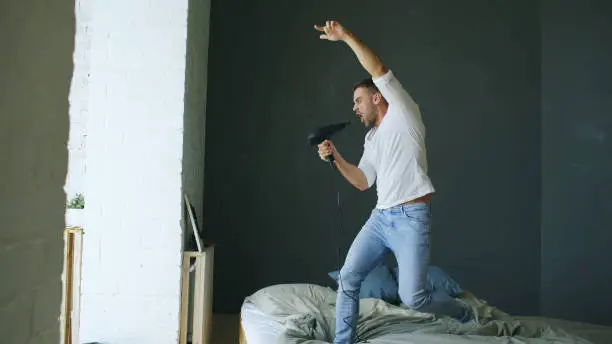 Photo of Young man singing to hair dryer and dancing rocknroll on bed in bedroom