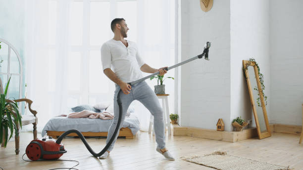 young man having fun cleaning house with vacuum cleaner dancing like guitarist - spring cleaning women cleaning dancing imagens e fotografias de stock