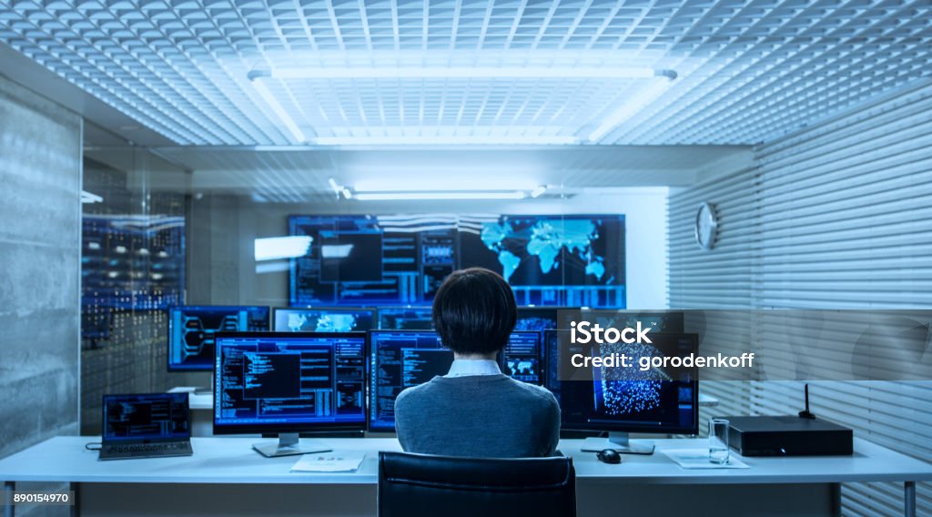 Back View of the IT Engineer Working with Multiple Monitors Showing Graphics, Functional Neural Network. He Works in a Technologically Advanced System Control Data Center. Network Security Stock Photo