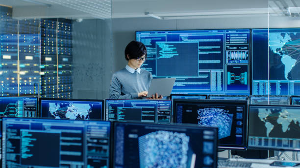 istock In the System Control Room IT Technician Holds and Works on a Laptop, in Background Multiple Displays with Graphics. Facility Works on Artificial Intelligence, Big Data Mining, Neural Network, Surveillance Project. 890155036