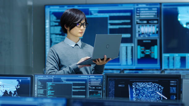 istock In the System Control Room IT Technician Holds and Works on a Laptop, in Background Multiple Displays with Graphics. Facility Works on Artificial Intelligence, Big Data Mining, Neural Network, Surveillance Project. 890155044