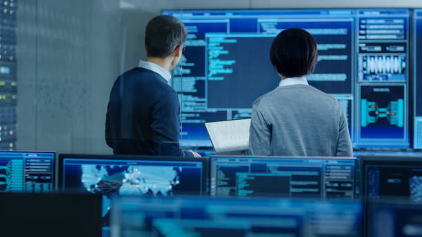 istock In the System Control Room IT Specialist and Project Engineer Have Discussion, they're surrounded by Multiple Monitors with Graphics. They Work in a Data Center on Data Mining, AI and Neural Networking. 890155054