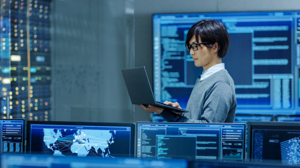 istock In the System Control Room IT Technician Works on a Laptop, in Background Multiple Displays with Graphics. Facility Works on Artificial Intelligence, Big Data Mining, Neural Network, Surveillance Project. 890155062