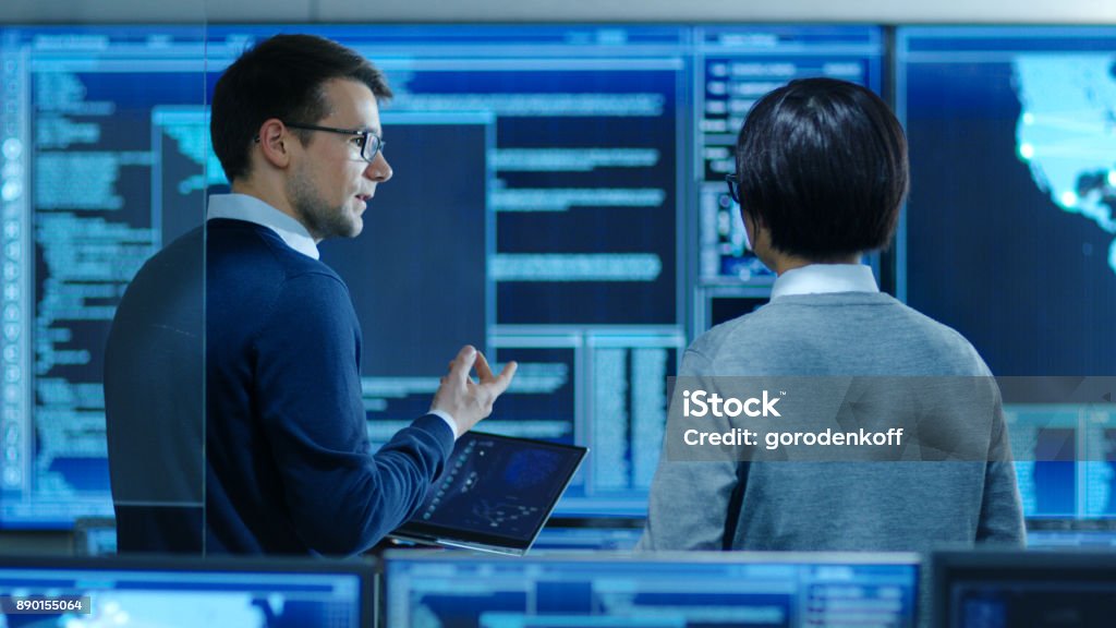 In the System Control Room IT Specialist and Project Engineer Have Discussion while Holding Laptop, they're surrounded by Multiple Monitors with Graphics. They Work in a Data Center on Data Mining, AI and Neural Networking. Network Security Stock Photo