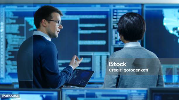 In The System Control Room It Specialist And Project Engineer Have Discussion While Holding Laptop Theyre Surrounded By Multiple Monitors With Graphics They Work In A Data Center On Data Mining Ai And Neural Networking Stock Photo - Download Image Now