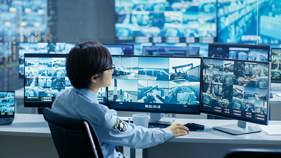 In the Security Control Room Officer Monitors Multiple Screens for Suspicious Activities, He Drinks from a Mug. He's Surrounded by Monitors and Guards Port of National Importance.