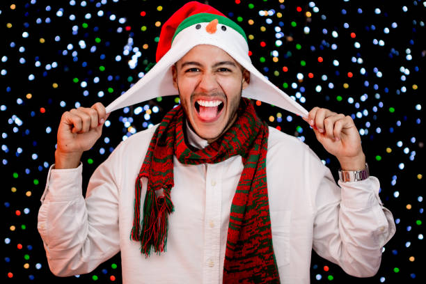 Young man poses wearing holiday scarf and hood. He smiles as pulling the hood with happiness for Christmas, Decoration lights for Christmas holiday in the bakground serve for setting the december mood.