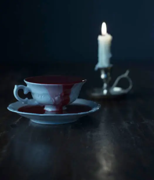 White tea cup in dark blood on dark wooden table with burning candle in candleholder.