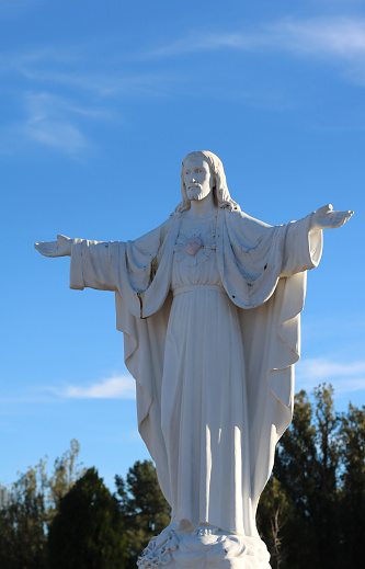 old statue of Jesus in stone against a bright blue sky, holding arms outward in a welcoming position, scars in palms, blue eyes and human heart around his neck in faded red