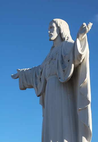 closeup of a stone Jesus statue in a cemetery, with arms outstretched backed by a bright blue sky