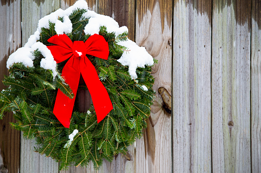 A Christmas pine wreath background with red bow hung in a winter snow storm on rustic barn wood with copy space.