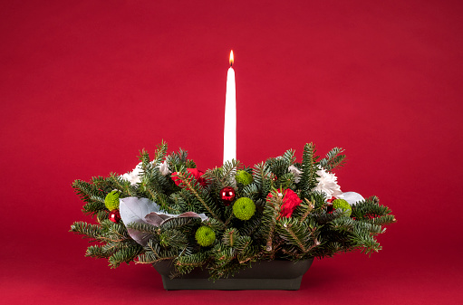 Christmas Table Arrangement or Centerpiece with flowers, evergreen branches and White Lit Candle on Red Background