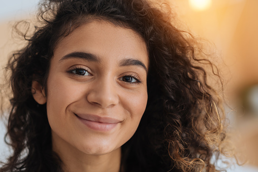 Heavenly beauty. Charming curly haired young lady looking into the camera with eyes full of happiness and smiling cheerfully indoors.