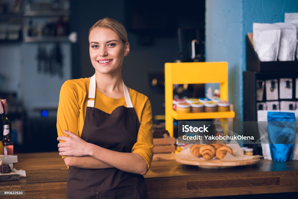 Smiling female worker situating in cafe Portrait of happy young barista standing near table while having job in confectionary shop. Job concept Retail Occupation Stock Photo