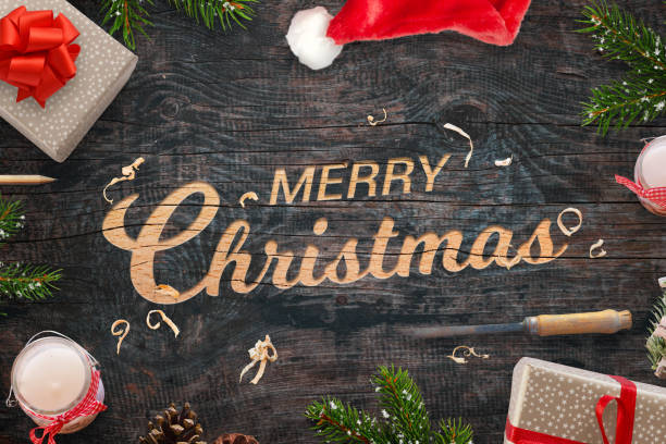 istock Merry Christmas greeting carved into a wooden surface. Christmas gifts, tree branches, chisel, candles, pinecones and Santa Claus hat beside. 890632524