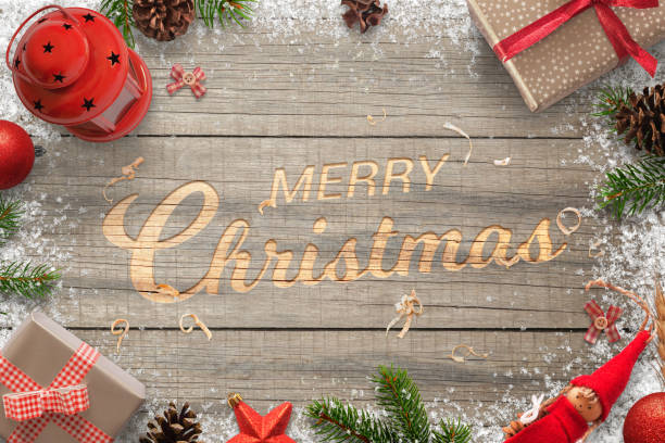 istock Creative hand carved Merry Christmas text in a wooden surface. Surrounded with Christmas decorations. Top view of branches, gifts, balls, lantern, pinecones and doll. 890632528