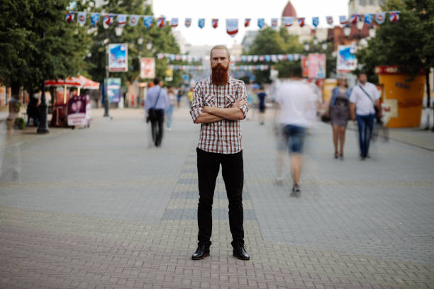 Young bearded man standing still at sidewalk in crowd traffic with people moving around outdoors