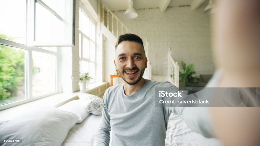 Young bearded man using tablet computer having video chat sitting in bed at home Young bearded hipster man using tablet computer having video chat sitting in bed at home Discussion Stock Photo