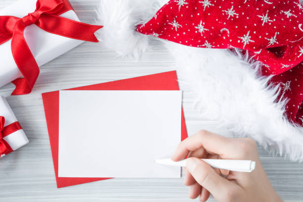 istock Woman's hand writing on the white blank greeting card. Santa Claus red hat and white gift boxes with ribbons on the cold, frosty wooden table. Christmas time concept. Empty place for a text. 890672036