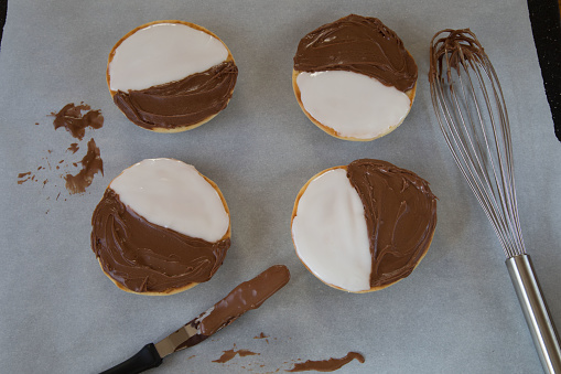 Four black and white cookies on a parchment paper surface with a whisk and offset spatula covered in chocolate.