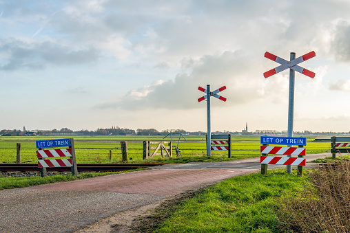 Backlit imnage of an unguarded level crossing in the Netherlands. On the blue signs with white letters you can see 'Caution train' (in the Dutch language).