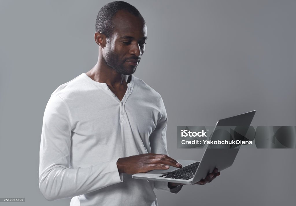 Skillful guy is typing on notebook Involved in working process. Waist-up of serious qualified young african businessman is standing and typing on modern laptop with concentration. Isolated background Adult Stock Photo