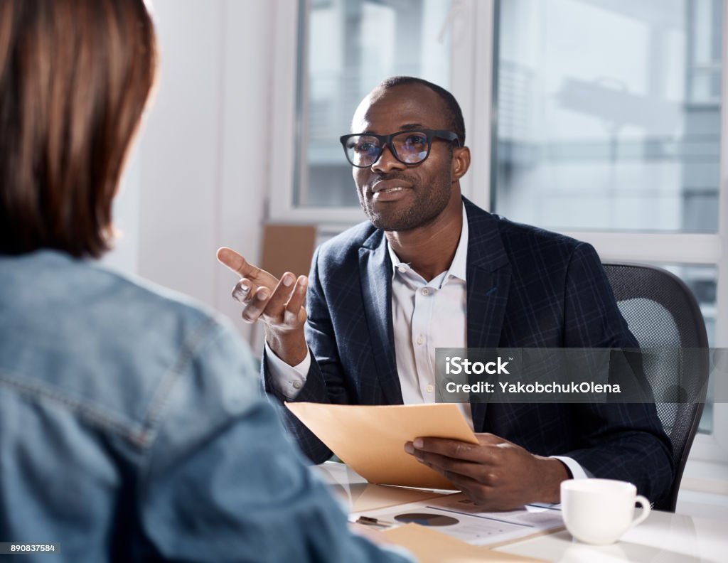 Skillful coworkers are discussing important project I listen to your suggestions. Portrait of confident qualified young african manager is sitting with his colleague female at table. He is looking at her attentively and gesticulating Interview - Event Stock Photo