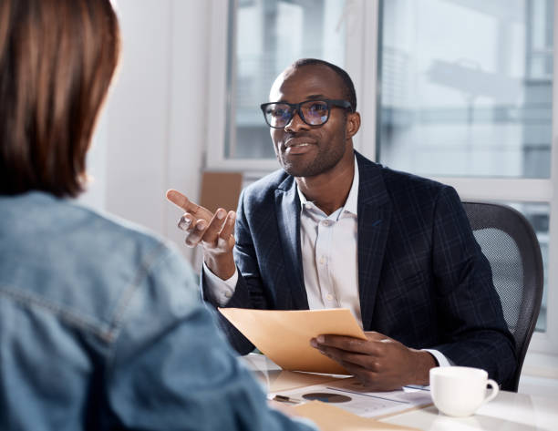 Skillful coworkers are discussing important project I listen to your suggestions. Portrait of confident qualified young african manager is sitting with his colleague female at table. He is looking at her attentively and gesticulating gesturing stock pictures, royalty-free photos & images