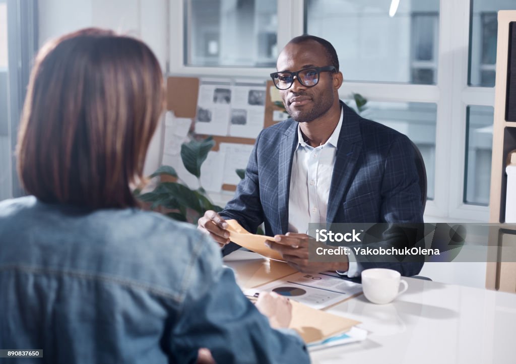 Professional colleagues are discussing project Listen attentively. Skillful young pleasant african businessman in suit is sitting at table and holding papers while looking at responsible colleague female who sitting in front of him Benefits Stock Photo