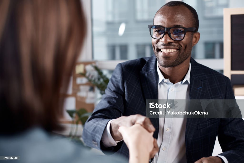 Optimistic qualified man is interviewing lady Nice to meet you. Portrait of cheerful young african manager in glasses is sitting at table and having firm handshake with new female employee. He is expressing gladness while looking at woman Job Interview Stock Photo