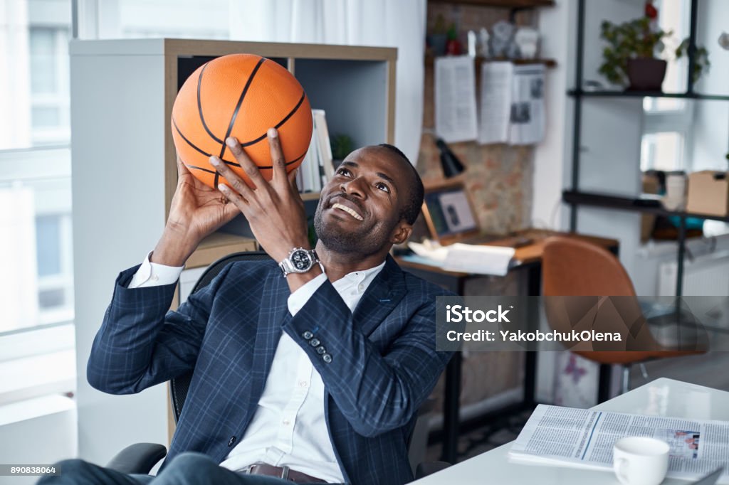 Joyful businessman is keeping ball in hands Playful mood. Cheerful young african manager in suit is sitting in office and holding orange basketball with smile. He is looking up while being ready to throwing it Manager Stock Photo