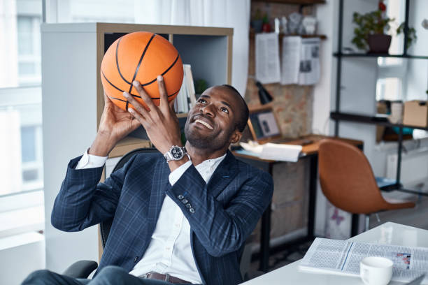 Joyful businessman is keeping ball in hands Playful mood. Cheerful young african manager in suit is sitting in office and holding orange basketball with smile. He is looking up while being ready to throwing it office competition stock pictures, royalty-free photos & images