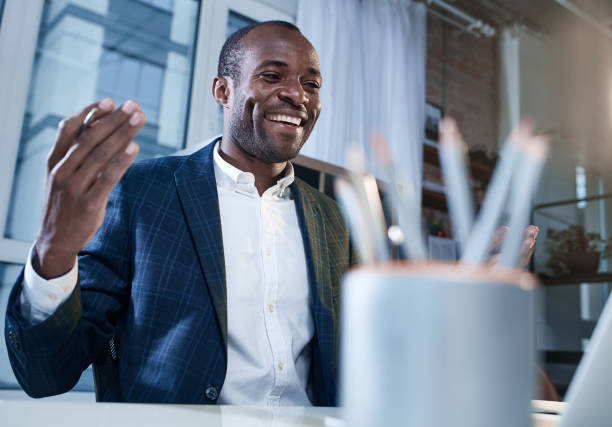 Joyful qualified man is feeling gladness Finally I did it. Low angle of cheerful young smart african businessman is sitting at table and looking at monitor of laptop while expressing happiness. Selective focus. Box with pencils on foreground qualified report stock pictures, royalty-free photos & images