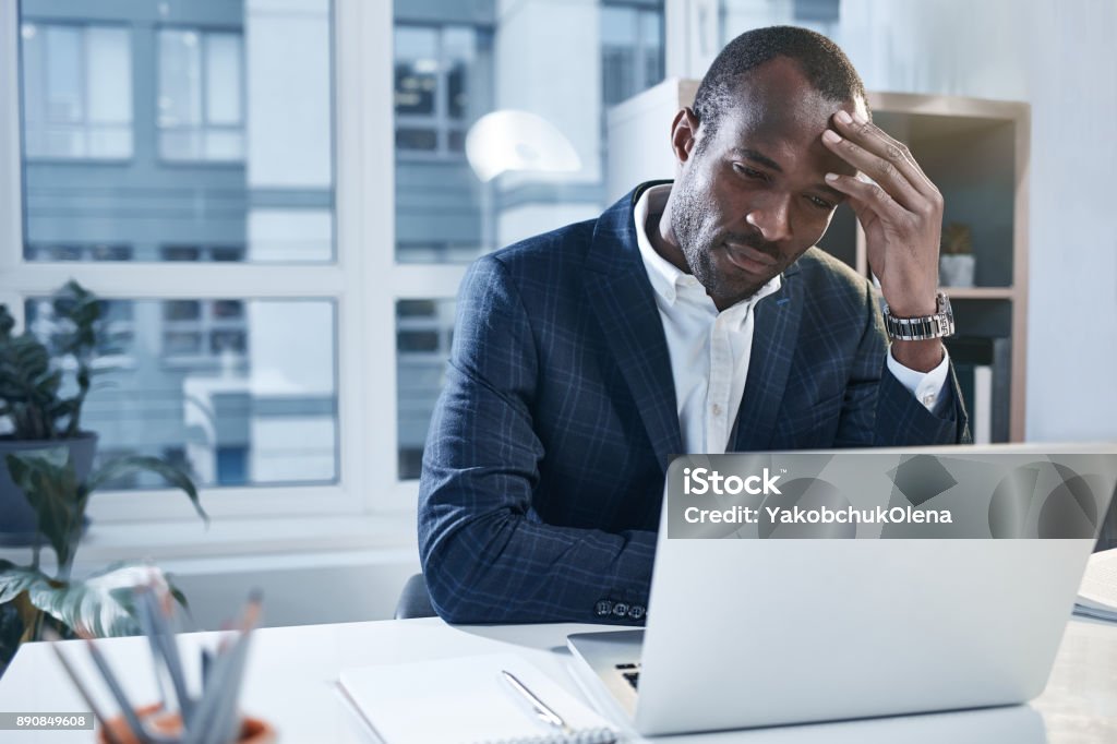 Exhausted pensive employee is using smart gadget Feeling headache. Tired young serious african businessman is sitting at desk and touching his head while expressing weariness. He is looking at screen of modern laptop with concentration. Copy space Businessman Stock Photo