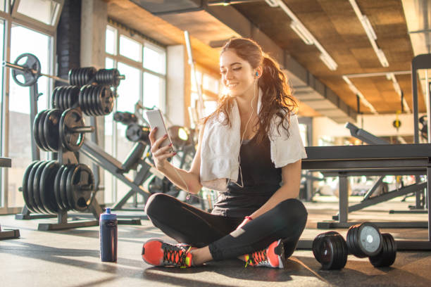 istock Smiling sporty girl with earphones using phone while sitting down on floor after hard workout in the gym. 890874246