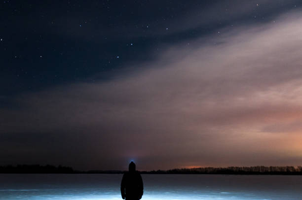 istock A man with a flashlight on his head is standing and looking at the beautiful starry sky with clouds. In a snowy field. Winter, a frosty night. 890935468