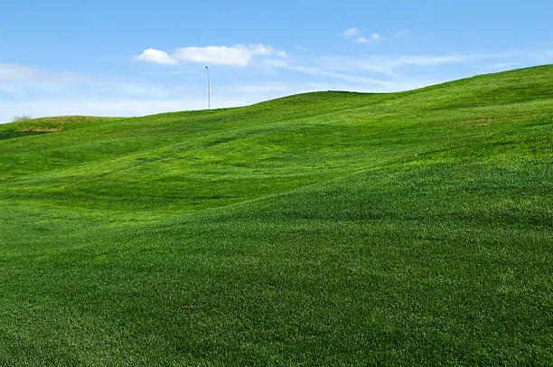 this color image is of rolling hills of green grass or lawn or a golf course fairway. the green grass has been mowed. and in the background is blue sky with white clouds. the photo was taken during the summer or spring. and the lighting is warm natural sunlight during the day or evening. the photo is a natural abstract background. 