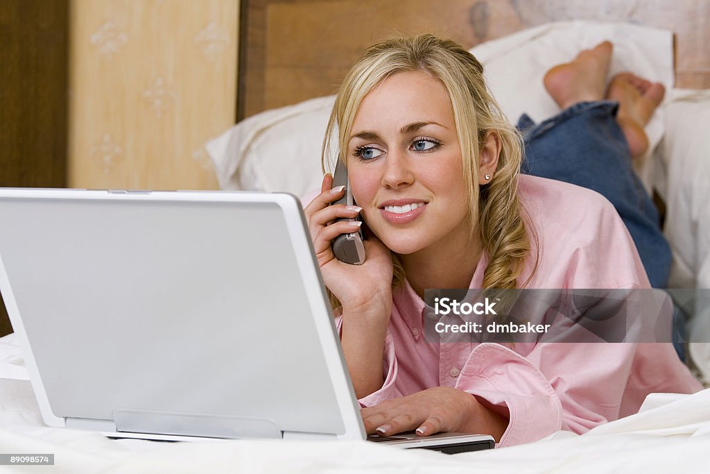Молодая женщина, лежа на кровати с телефона и ноутбука - Стоковые фото Беспроводная технология роялти-фри