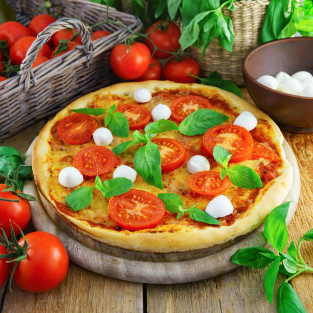 istock Italian pizza with mozzarella, cheese, and basil leaves on rough rustic rural planks table. Wicker wooden basket with fresh raw organic tomatoes. Square, selective focus. 890994362