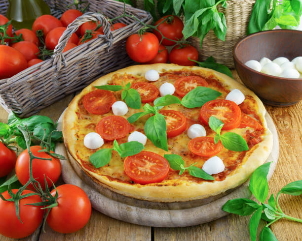 istock Italian pizza with mozzarella, cheese, and basil leaves on rough rustic rural planks table. Wicker wooden basket with fresh raw organic tomatoes. Horizontal, selective focus. 890994374