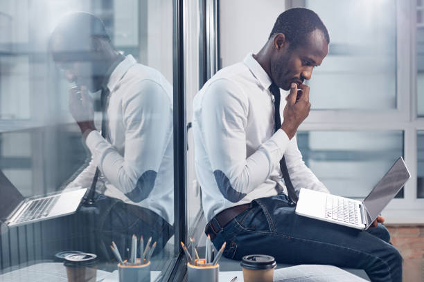 Pensive stylish businessman is making important task Effective planning. Side view profile of african man is sitting on windowsill while leaning on glass and holding laptop on knees. He is looking at screen of gadget thoughtfully while touching his chin introspection photos stock pictures, royalty-free photos & images