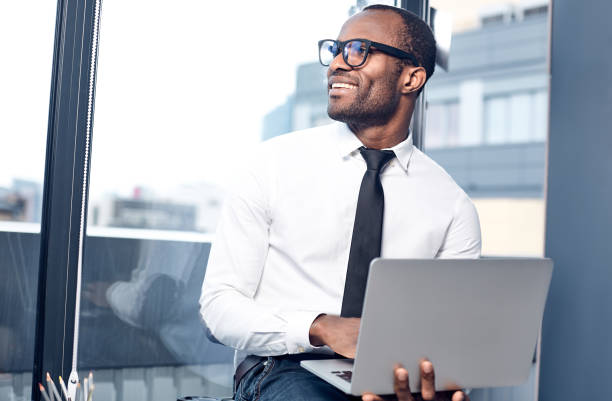 Cheerful qualified manager is typing on notebook Pleasant atmosphere. Joyful elegant young african businessman in glasses is sitting on desk and looking aside with smile while holding modern laptop. Big window in background qualified report stock pictures, royalty-free photos & images