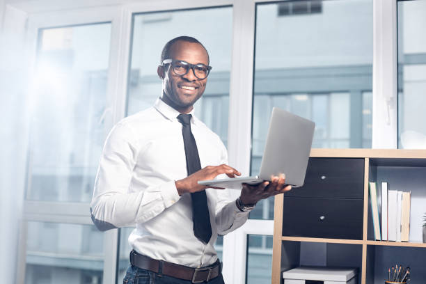Optimistic qualified man is using modern device Enjoying my job. Portrait of positive young elegant african businessman is holding laptop and typing on it while standing in office and looking at camera with joy. Big window in background qualified report stock pictures, royalty-free photos & images