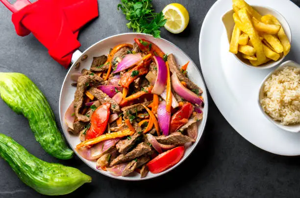 Peruvian dish Lomo saltado - beef tenderloin with purple onion, yellow chili, tomatoes in pan, fried potatoes french fries and rice on slate background. Tot view