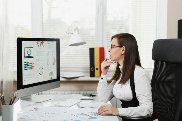 istock Beautiful business woman in suit and glasses working at computer with documents in light office, looking at the monitor 891265460