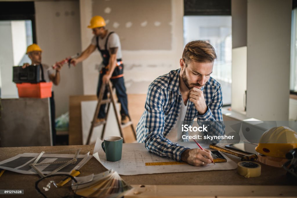 Male architect drawing improvements on housing plan at construction site. Pensive architect drawing improvements on housing project inside of a built structure. Workers are in the background. Apartment Stock Photo