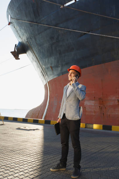 istock Handsome young unshaven successful business man in gray suit and protective construction orange helmet holding tablet, talking on mobile phone, standing in sea port against cargo rusty ship background 891297380