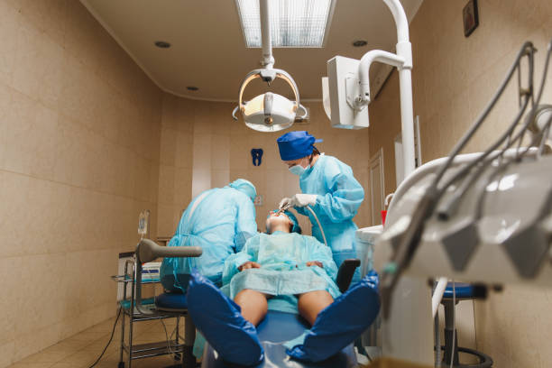 制服と女性アシスタントの男性の古いプロの歯科医師の外科医は、現代のツール装備クリニック光オフィスで女性患者の操作インストール歯科インプラント歯を実行するのに役立ちます。 - dental light dental equipment hospital professional occupation ストックフォトと画像