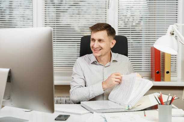 istock Handsome smiling business man in shirt sitting at the desk, working at computer, black folder, documents in light office, looking to contemporary monitor, on window background. Manager or worker. 891307090