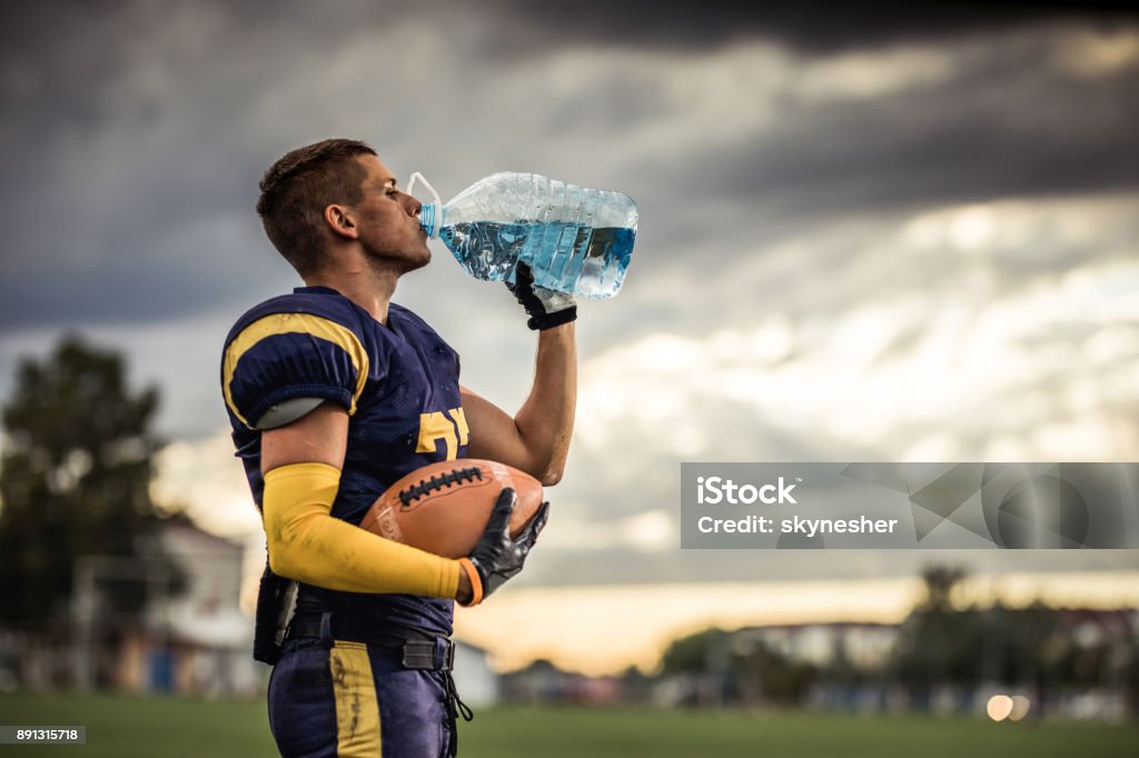 Thirsty American football player drinking fresh water on playing field. Profile view of young American football player drinking water on a break at playing field. Drinking Stock Photo